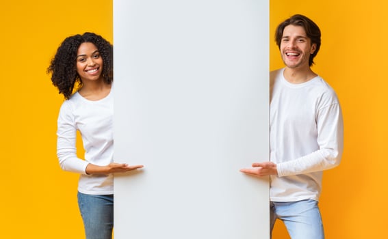 Cheerful Guy And Afro Girl Holding And Pointing At White Advertisement Board, Standing Together Over Yellow Background photo