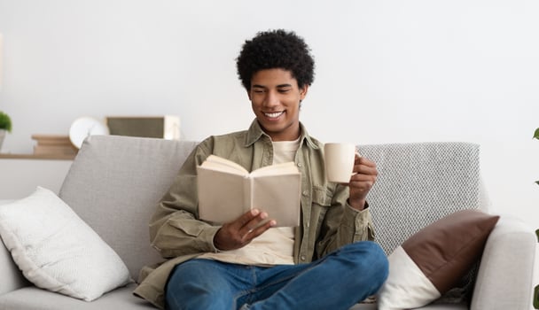 Positive black teenager reading book and drinking coffee while sitting on sofa at home. Smiling African American teen guy studying remotely from home, making assignment, looking through textbook photo