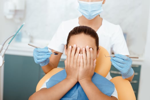 Kids fear of medical instruments. Scared little latin girl in dental chair covers face and does not give young woman doctor in white coat, protective mask and gloves examination teeth in clinic photo
