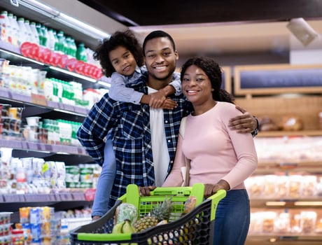 Portrait of positive black family with shopping cart smiling and looking at camera at modern supermarket. Young parents and their cute little daughter buying groceries at huge mall photo