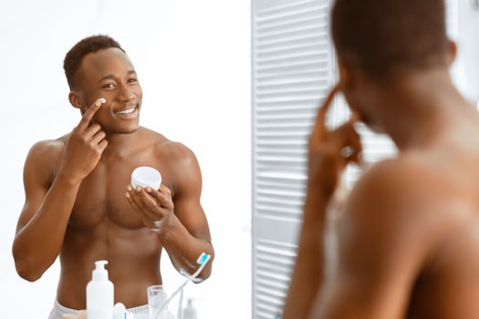 Facial Skincare Concept. Handsome Black Guy Applying Moisturizer Cream On Face Standing Shirtless Near Mirror In Modern Bathroom At Home. Male Skin Care Routine. Selective Focus photo