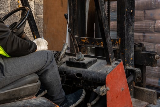Close up of a forklift operator in black jacket and gloves using controls. Worn metal parts, levers, and packaged goods visible near a textured wall. photo