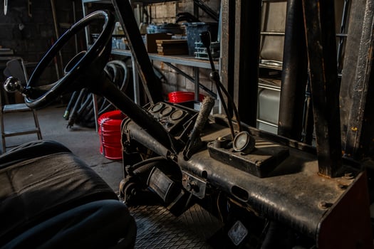 Weathered forklift interior showing steering wheel, control levers, and analog gauges. Industrial tools, hoses, and shelving visible in the setting. photo