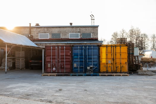 Red, blue, and yellow shipping containers on pallets near a brick industrial building with windows, a chimney, and scattered materials in warm light. photo