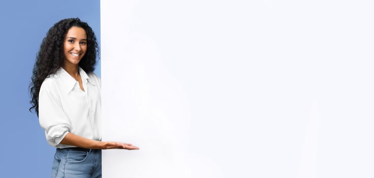Place For Your Ad. Smiling brunette woman pointing at white advertisement board with open palm, demonstrating copy space for your text or design, standing over yellow studio background, panorama photo