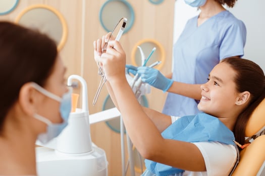 Satisfied dentist clinic patient look at her perfect smile in mirror after treatment in office with medical equipment in background and nurse in white coat, protective mask and rubber gloves, cropped photo