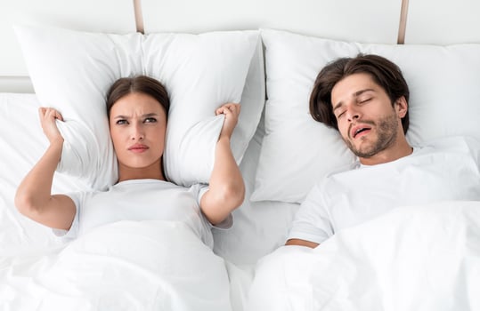 Attractive millennial european guy with stubble sleeps on bed, angry upset wife covers ears with pillow, suffers noise in bedroom interior. Snoring, health and relationship problems, apnea, insomnia photo