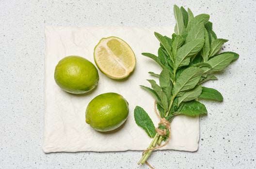 Whole limes and a halved lime are arranged next to a small bunch of fresh mint leaves tied with twine photo