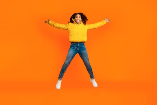 Joyful African American Girl Doing Spread Eagle Jump Posing Wearing Winter Jacket Smiling To Camera Over Yellow Studio Background photo