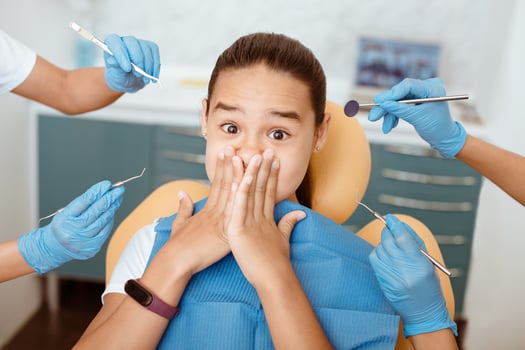 Children fear, emotions of horror and treatment of oral cavity in clinic. Scared girl with wide open eyes covers her mouth with her hands in chair, around hands of doctors in rubber gloves with tools photo