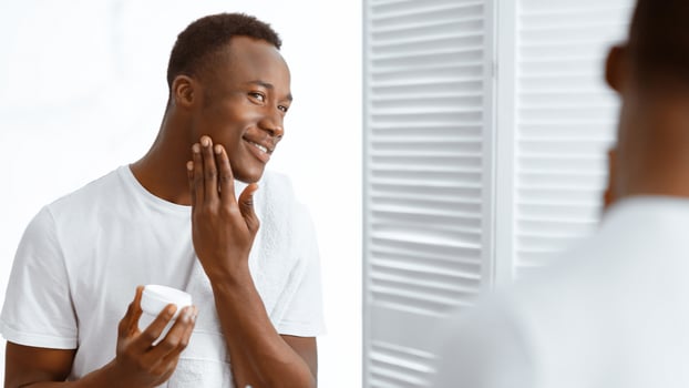 Male Facial Skincare. Black Man Using Moisturizing Aftershave Cream Caring For Face Skin Standing Near Mirror In Bathroom At Home. Men's Beauty And Self-Care Routine Concept. Panorama photo