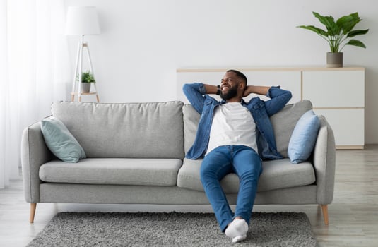 Cheerful attractive millennial african american male with beard sitting on couch and look at empty space in minimalist living room interior. Rest at home, break. Man enjoys weekend and free time photo