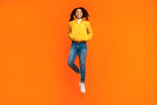 Joyful African American Woman Jumping Posing Wearing Winter Jacket Over Yellow Studio Background, Smiling To Camera. Female Jump. Full Length Shot, Empty Space For Text photo
