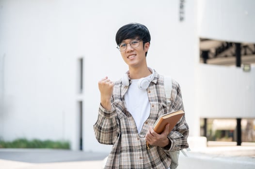 An asian man with glasses is holding books and making a fist while standing in front of a building. photo