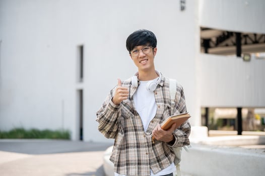 An asian man with glasses is holding books and making thumbs up while standing in front of a building photo