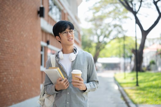 A smiling asian man with glasses is holding books with coffee cup while standing near brick wall. photo