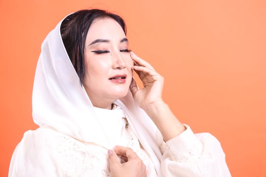A contemplative Asian woman wearing a white hijab and elegant makeup with her eyes closed, lost in thought or prayer isolated on a orange background. photo