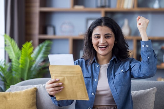 An excited young woman sitting on a couch with a letter in her hand, looking overjoyed, raising her fist in the air in celebration. photo