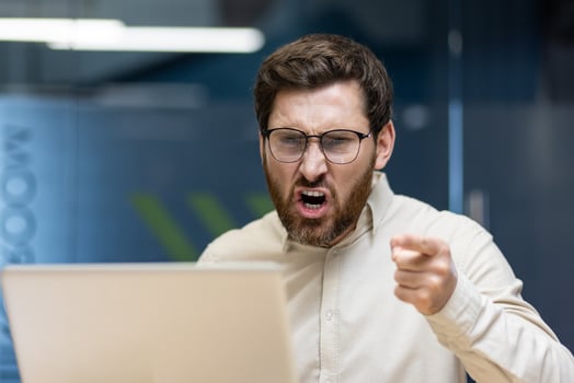 Close-up photo of a young angry man sitting in the office at the desk and shouting while pointing his finger at the laptop screen.