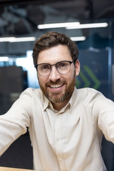 Vertical close-up photo of a young man in glasses and a shirt who is in the office and prays to the online camera he is holding in his hands.