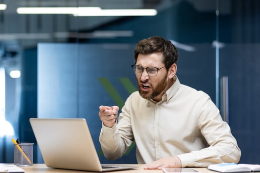 Angry young businessman sitting in the office at the desk, talking aggressively, arguing over a call, pointing a finger at the laptop screen. photo