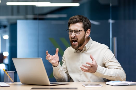 Angry young male office worker sitting at workplace and frustratedly yelling at laptop screen, gesturing with hands. photo