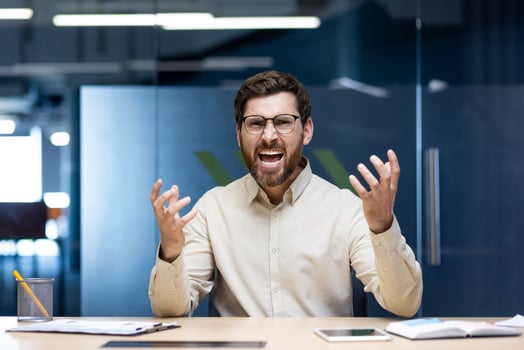 Close-up portrait of an angry and irritated young man sitting in an office at a desk and yelling at the camera while waving his hands. photo