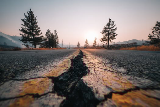 A deep crack cuts through a forest road at golden hour with pine trees and mountain silhouettes in the background. Perfect for themes of seismic damage, isolation, or nature reclaiming infrastructure. photo