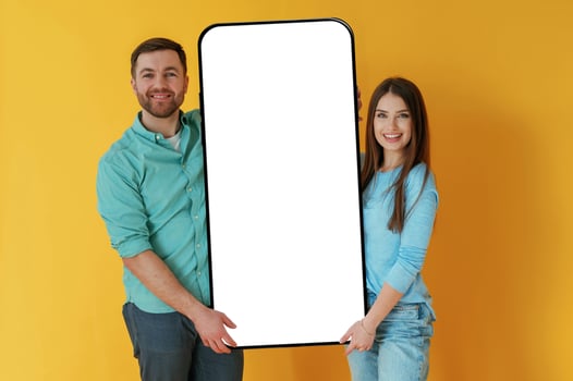 Holding, white display of smartphone, copy space. Man and woman are standing against yellow background photo