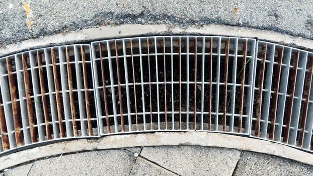 Curved drainage channel blocked by steel plates. Side of the footpath is made of concrete slabs. and asphalt road outside. photo