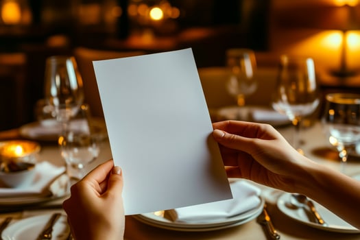 Hands holding a blank white menu card with an elegant dining table setup, glowing candlelight, and refined glassware in a cozy restaurant ambiance photo