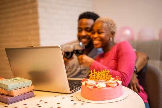 Happy African couple celebrating birthday together at home, smiling and toasting with wine glasses over birthday cake during online call on laptop. Selective focus on cake. photo