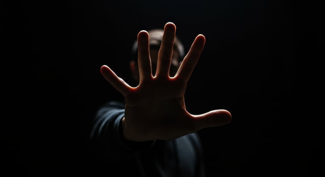 Man raises hand in front of face in dark setting, fingers spread. Gesture of stopping, blocking, or Vulcan salute concept on black background. photo