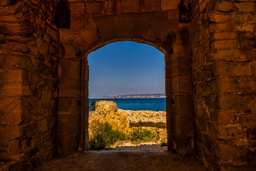 A stone archway frames a stunning view of the sea under a clear blue sky, with rocky terrain leading to the shoreline photo
