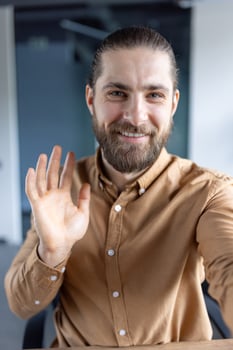 A bearded man waves at the camera, smiling. He wears a brown shirt and seems to be in a professional setting, possibly an office. photo