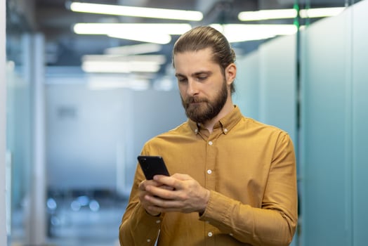 A man with a beard and his hair tied back is looking at his phone in a corporate office setting. photo
