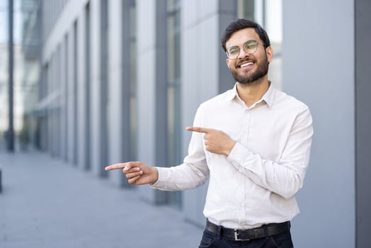 A smiling man with glasses stands outside, pointing towards something. He is wearing a white shirt and looking directly at the viewer. The background has a modern building. photo