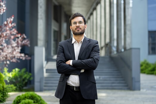 A well-dressed man stands confidently outdoors, arms crossed, looking directly at the camera with a serious expression. The backdrop features a modern building. photo