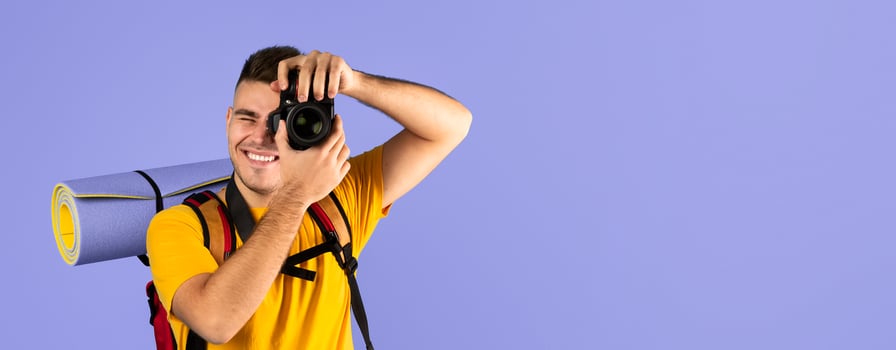 Happy millennial guy with tourist equipment taking photo on professional camera over blue studio background. Smiling male photographer shooting picture during his camping trip