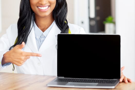 Positive female nutrition specialist pointing at laptop with blank screen at medical office, mockup for your website. Professional black dietitian promoting weight loss clinic, space for design photo