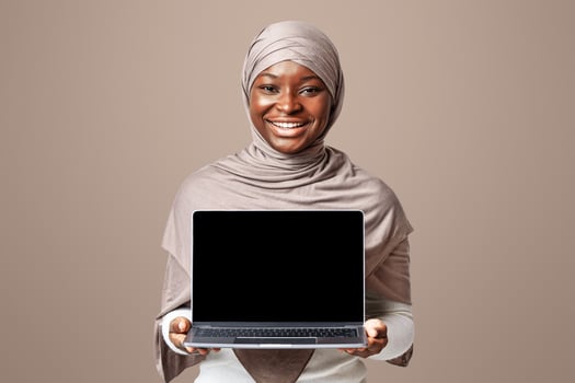 Great Website. Cheerful African Muslim Woman In Hijab Holding Laptop Computer With Blank Black Screen, Demonstrating Copy Space For Your Design, Standing Over Light Studio Background, Mockup Image photo
