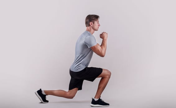 Side view of young fit guy in sportswear doing lunge on light studio background. Professional millennial bodybuilder warming out before strength training, doing aerobic exercises photo