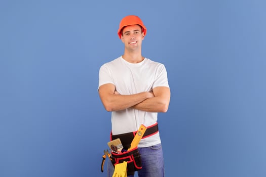 Confident young couple in hard hats ready for house repairment and renovation, millennial spouses in casual clothes standing with folded arms over blue background, man wearing tool belt, copy space photo