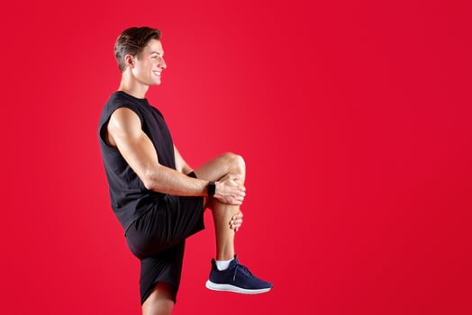 Full length portrait of sporty Caucasian man stretching his leg over red studio background. Fit millennial guy in sports clothes warming up before workout, doing flexibility exercises photo
