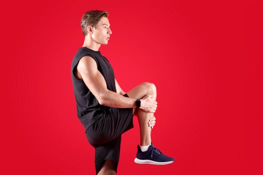 Flexible millennial man stretching his leg over red studio background, side view. Athletic young guy in sportswear warming up before training, practicing flexibility exercises photo