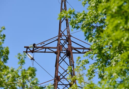 Upper part of an old, rusty power line tower against a blue sky and green tree leaves. photo