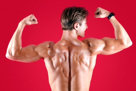 Young muscular sportsman standing with his naked back to camera and showing biceps on red studio background. Handsome athletic guy posing and demonstrating his strong muscles photo
