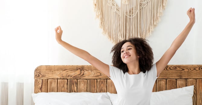 Good morning, new plans for day, dreams, great mood and vitality. Happy millennial cute african american woman sitting on white comfortable bed and stretching, in cozy bedroom interior, free space photo