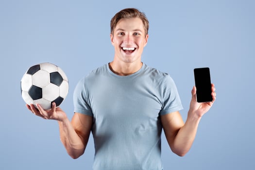 Happy young guy holding soccer ball and smartphone with blank screen on lilac studio background, mockup for design. Positive football fan showing mobile device with space for website or app photo