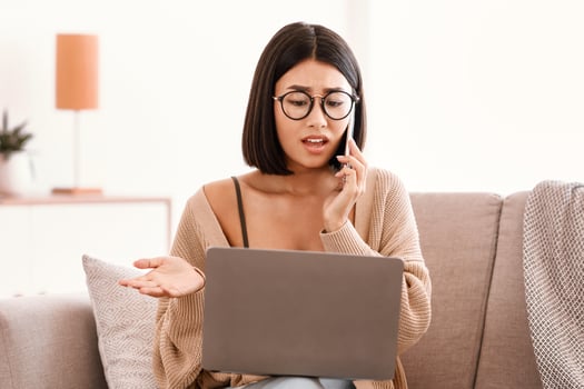 Problem Concept. Frustrated serious woman in glasses talking on mobile phone, sitting on couch and using laptop, having problem with internet connection, calling customer support hotline photo